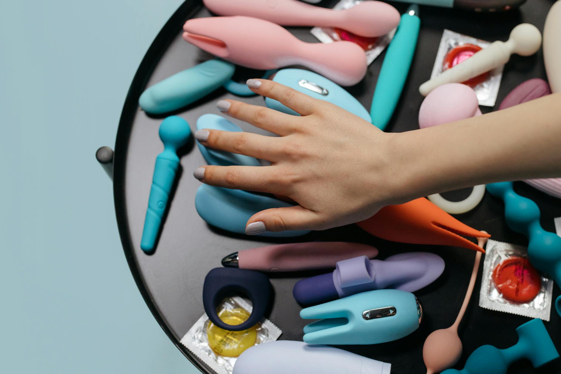 A hand reaching over a variety of colorful sex toys, including suction toys and vibrators, arranged on a table.