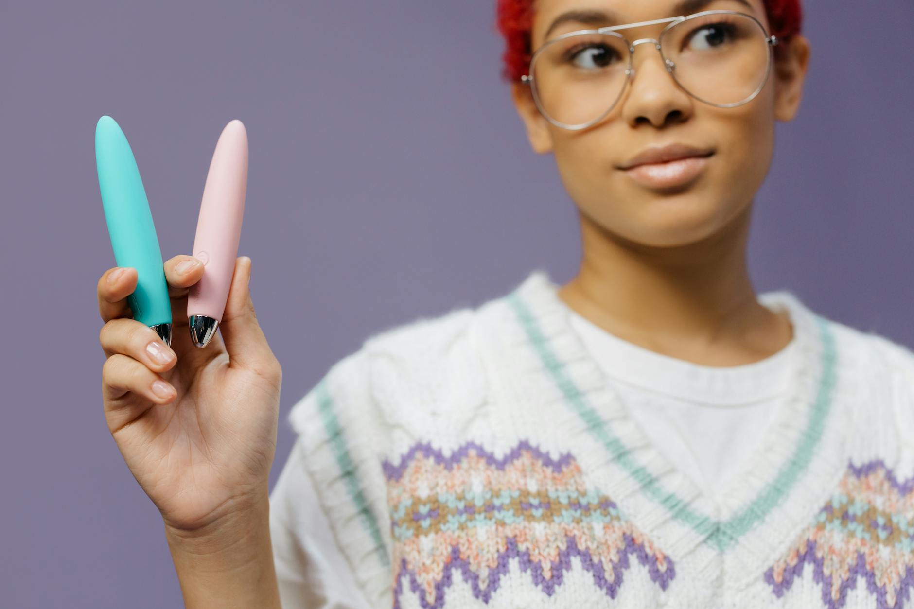 Una giovane donna con occhiali tiene due vibratori colorati su uno sfondo viola.